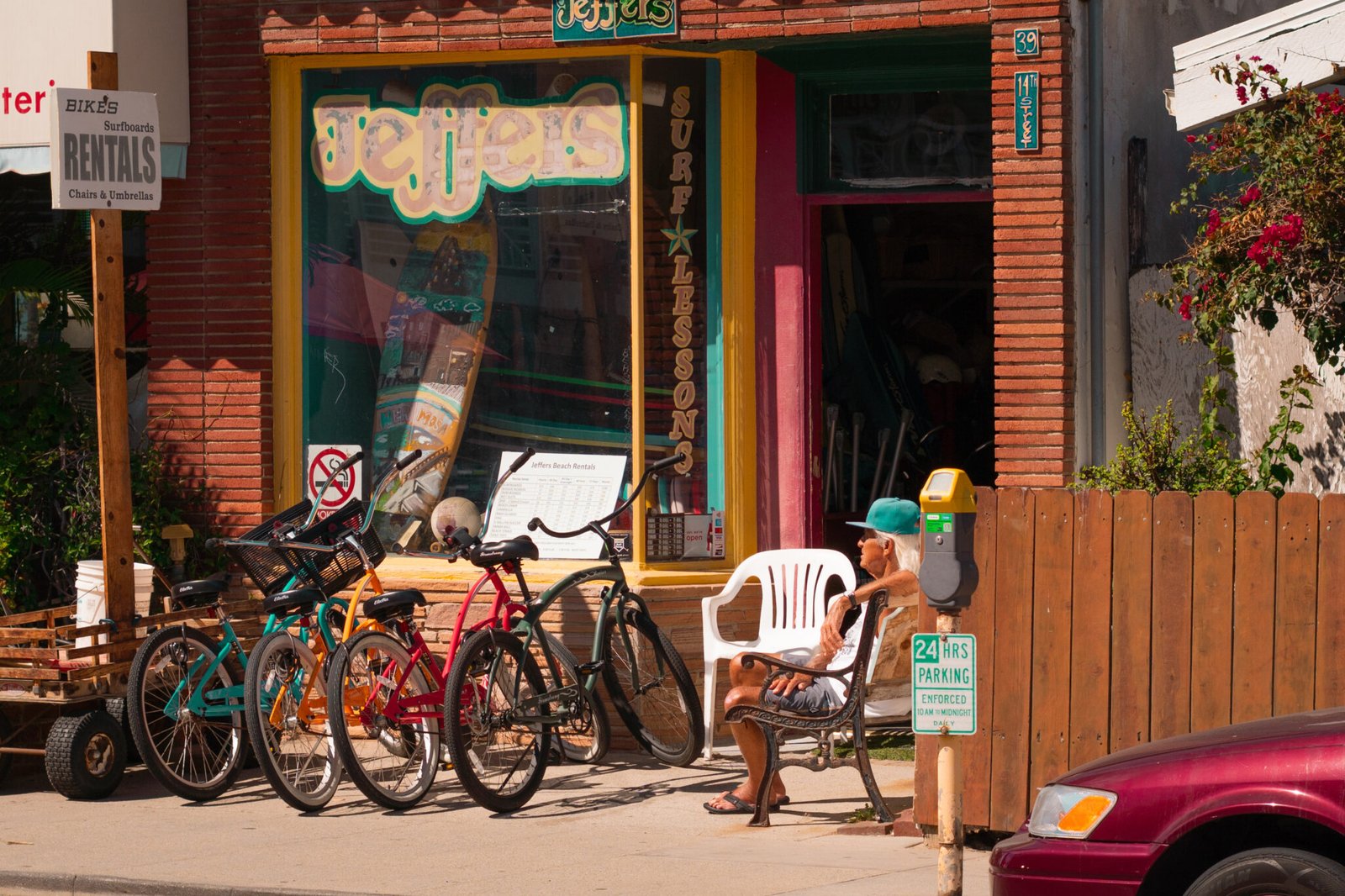Overlooking "Jeffers" surf shop, an older local with a blue hat on sits on the bench enjoying the warm day. In front of him are four bikes; yellow, blue, red, and dark green.