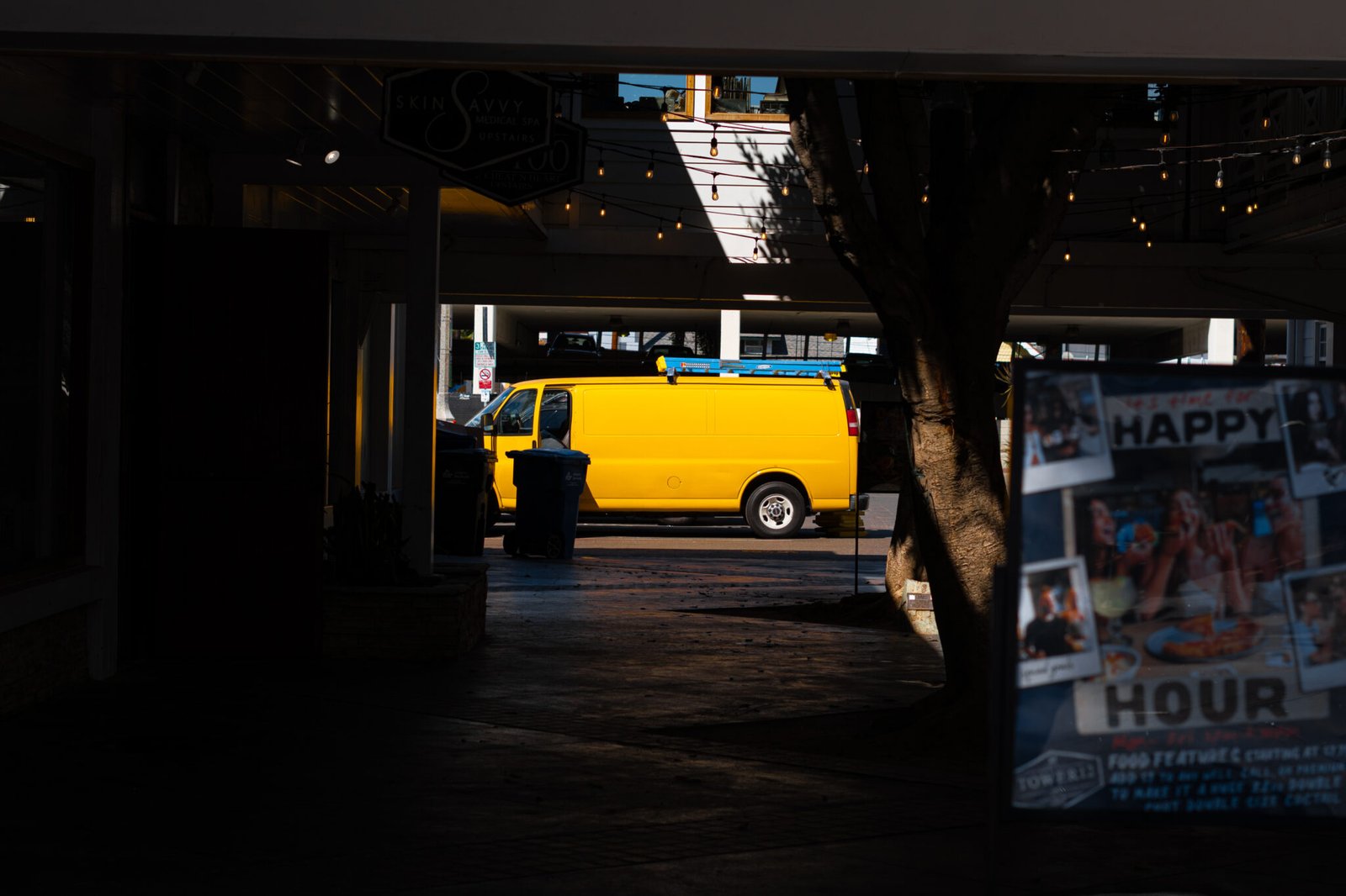 The picture is taken from underneath a dark overpass. On the other side of the overpass is a bright street. On the street is a bright yellow/orange worker van with a light blue ladder on its roof. The yellow light from the van bounces within the dark overpass, creating some bright reflections.